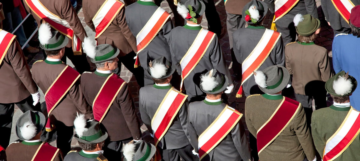 The picture shows a group of people photographed from behind. They are wearing traditional uniforms with hats and diagonal sashes in different colors.
